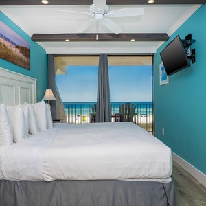 This bedroom features a plush bed, vibrant blue walls, and a balcony overlooking the beach, complemented by a wall-mounted TV
