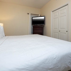 Spacious bedroom features a king bed with plush white linens and a TV mounted above a dark wood dresser