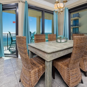 A dining area features a rustic wooden table surrounded by woven chairs, with large windows offering a beach front view