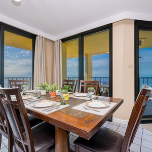 A wooden dining table set for six offers a view of the beach front through floor-to-ceiling windows