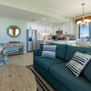 Living area features a teal sofa with striped pillows, a rustic coffee table, and a dining space with a view of the balcony