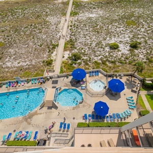 Aerial view of the pool area featuring a large swimming pool, kiddie pool, and lounge chairs under blue umbrellas