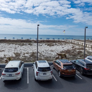 A view of the beach with blue umbrellas and a wooden walkway, framed by parked cars in a well-maintained lot