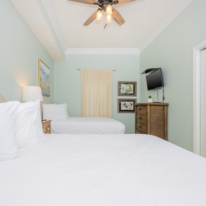 Bright guest bedroom featuring two double beds, decorative artwork, a flat-screen TV, and a charming wicker dresser