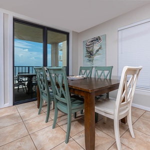 This dining area features a wooden table with teal and white chairs, with a stunning beach view from the balcony