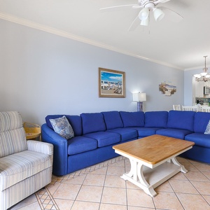 The living area features a vibrant blue sectional sofa, a wooden coffee table, and a view of the kitchen with bright cabinets