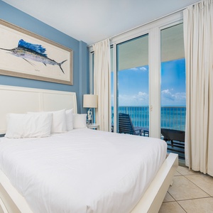 The guest bedroom features a plush white bed, a TV, and stunning beachfront views with balcony access