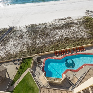Aerial balcony view showcasing the pool area with vibrant orange loungers, surrounded by lush grass and leading to a beach