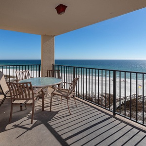 The balcony features a glass-top table with chairs, offering a serene view of the beach and shimmering water beyond