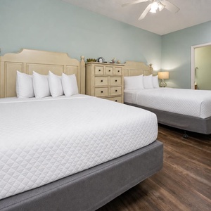 This guest room features two queen beds with plush white bedding, a light wood dresser, and soothing blue walls