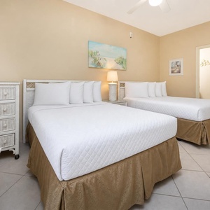 Bright guest room featuring two double beds, a wicker dresser, and coastal artwork, creating a relaxed atmosphere