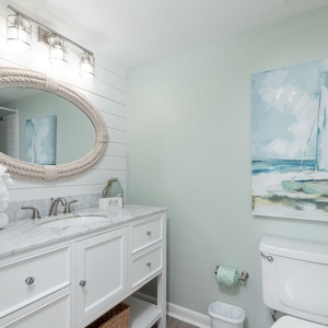The guest bathroom features a marbled countertop, a large oval mirror with a rope frame, and beach-themed artwork on the wall