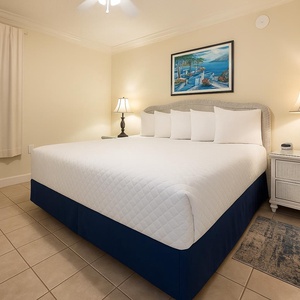 The bedroom features a plush king bed, bedside lamps, and a vibrant coastal painting above the headboard