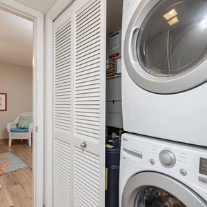 A laundry area features a stacked washer and dryer beside a cozy guest bedroom with colorful decor and a wicker chair