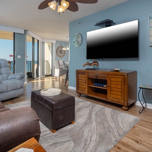 The living area features a cozy brown sofa, plush blue recliner, and a large TV, with a view of the beach