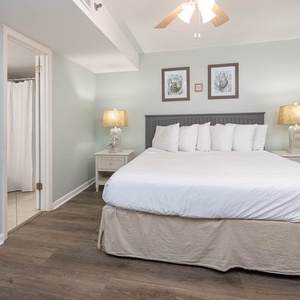 Serene bedroom features a king bed with plush pillows, soft lighting from bedside lamps, and coastal artwork on the walls
