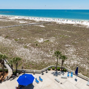 Expansive view of the beach with white sand and turquoise gulf waters, featuring a pool area and lounge chairs below
