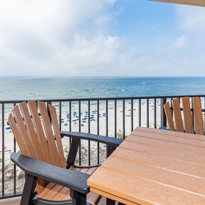 Enjoy a relaxing view from the balcony with wooden chairs overlooking the beach and vibrant blue umbrellas dotting the shore