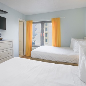 The bedroom features two comfortable beds, a TV, and bright curtains framing a view of the beach