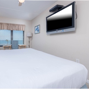 This inviting bedroom features a plush bed, a wall-mounted TV, and a cozy seating area by the window overlooking the gulf
