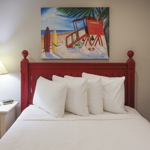 This guest bedroom features a vibrant red bed frame, bright white linens, and a cheerful beach-themed painting
