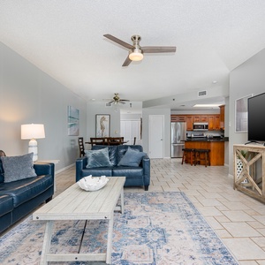 Living area featuring a dark blue sofa with queen sleeper, light wood coffee table, and a large TV, leading to the kitchen