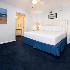 Master bedroom features a king bed with plush linens, a nightstand with a lamp, and a cozy blue carpet