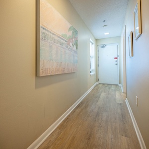 Welcoming hallway features light wood flooring, soft colors, and artwork, leading to a door and natural light from a window