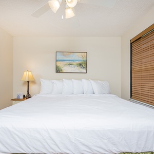 A guest bedroom features a king bed with multiple pillows, a bedside lamp, and a serene beach-themed painting on the wall