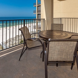 The balcony features a round table with four chairs, offering a direct view of the beach and gentle waves below