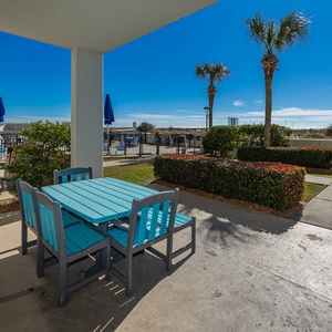 Outdoor patio area features a turquoise table and chairs, surrounded by lush greenery and views of the pool area
