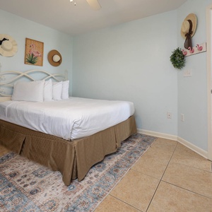 Guest bedroom features a queen bed with white linens, decorative wall art, and a glimpse of the kitchen beyond