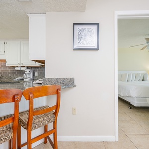 Kitchen with white cabinetry and a granite countertop, featuring two wooden bar stools and a glimpse of the bedroom beyond