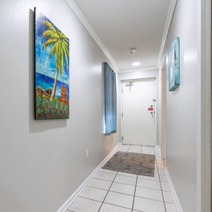 Colorful artwork adorns the walls of a bright hallway leading to the entrance, with soft curtains and tiled flooring