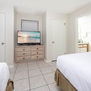 Bright guest room featuring two double beds, a TV on a wooden dresser, and a doorway leading to a well-lit bathroom area