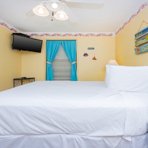 This cozy guest bedroom features a queen bed, bright yellow walls, a wall-mounted TV, and cheerful blue curtains