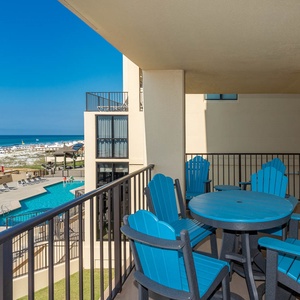 The balcony features a round table with blue Adirondack chairs and offers a clear view of the pool and beach front