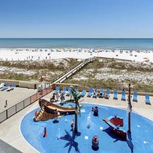 Vibrant pool area features a playful water play splash zone with a pirate ship, surrounded by lounge chairs and a beach view
