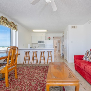 The living area features a cozy red sofa, a TV, and a dining table with a beautiful beachfront view