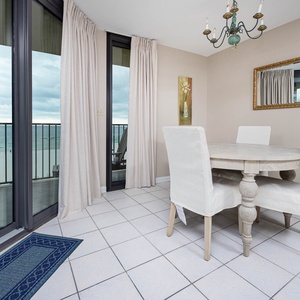 Light-filled dining area features a round wooden table with white upholstered chairs and a stunning beachfront view
