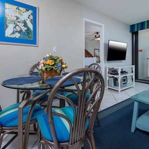 The living area features a glass dining table, colorful decor, and a TV, with a stunning beach view through the sliding doors