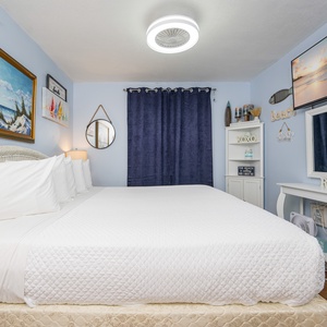 The bedroom features a cozy white bed adorned with plush pillows, beach-themed artwork, and soft lighting for a relaxing stay