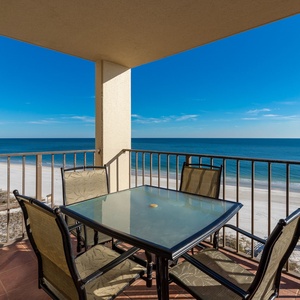 Balcony with a glass-top table surrounded by chairs, offering a panoramic view of the tranquil beach and clear blue water