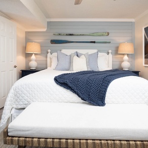The bedroom features a white quilted king bed with blue accents, stylish lamps, and a bathroom entrance visible nearby