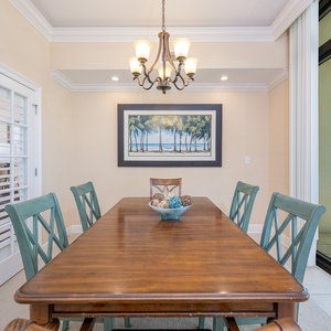 Dining area featuring a wooden table, coastal artwork, and large windows showcasing a serene beachfront view