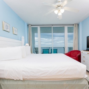 This bedroom features a king-size bed, a vibrant red chair, and a stunning beachfront view from the sliding glass doors