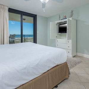 Brightly lit master suite featuring a plush king bed, a TV in a white cabinet, and a balcony with a view of the beach beyond