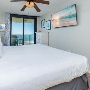 The master bedroom features a large king bed, a mounted TV, and sliding glass doors leading to a balcony with a beach view