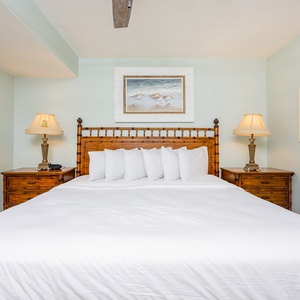 The bedroom features a king bed with plush white bedding, bedside lamps, and artwork depicting coastal scenes