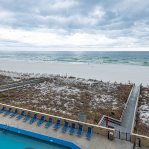 Tranquil beachfront with white sand, a pool area lined with blue loungers, and gentle waves in the distance
